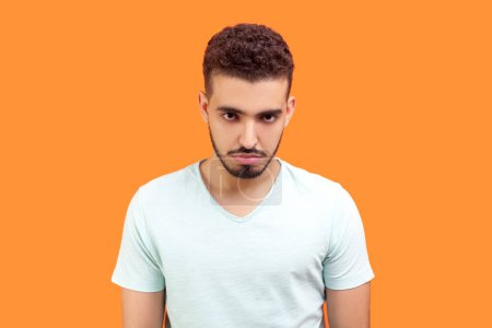 Photo for Portrait of sad unhappy young bearded man wearing T-shirt looking at camera with sad upset expression being in bad mood. Indoor studio shot isolated on orange background. - Royalty Free Image