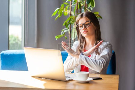 Photo for Portrait of serious strict woman working on laptop no way gesture crossed her arms looking with bossy expression, wearing jacket and re shirt. Indoor shot, cafe or office background. - Royalty Free Image