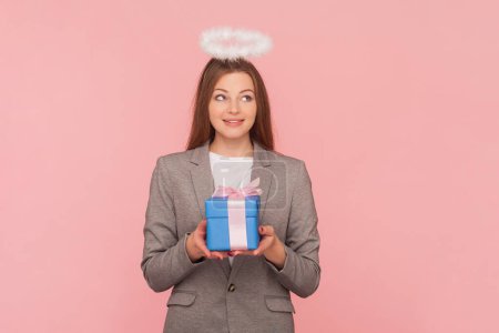 Photo for Portrait of dreaming smiling woman with brown hair with nimb over head holding gift box, making wish on birthday, wearing business suit. Indoor studio shot isolated on pink background. - Royalty Free Image