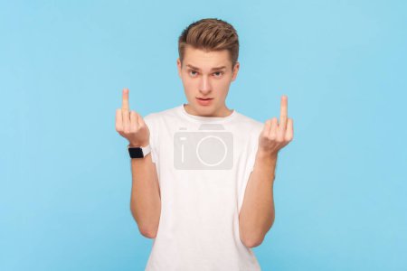Téléchargez les photos : Portrait of strict serious man wearing white t-shirt showing middle finger fuck sign, arguing with somebody, being rude and impolite. Indoor studio shot isolated on blue background - en image libre de droit