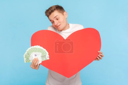 Photo for Portrait of rich delighted man wearing white t-shirt holding big red heart and fan of euro banknotes, love to money. Indoor studio shot isolated on blue background - Royalty Free Image