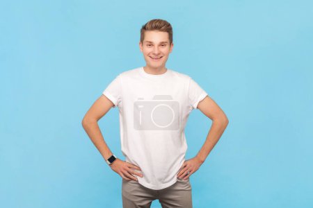 Photo for Portrait of confident cheerful smiling man wearing white t-shirt standing with hands on hips, looking at camera, being extremely joyful. Indoor studio shot isolated on blue background - Royalty Free Image
