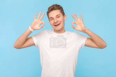 Photo for Well done. Portrait of delighted attractive blonde man wearing white t-shirt showing okay gesture, approved work, being satisfied. Indoor studio shot isolated on blue background - Royalty Free Image