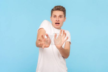 Photo for Portrait of scared frighten blonde man wearing white t-shirt outstretching hands, trying to avoid danger, looking with panic at camera. Indoor studio shot isolated on blue background - Royalty Free Image