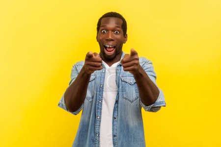 Photo for Portrait of amazed excited surprised man pointing at camera choosing you expressing optimistic emotions, wearing denim casual shirt. Indoor studio shot isolated on yellow background. - Royalty Free Image