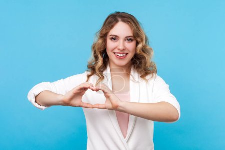 Photo for Portrait of lovely cute blond woman with wavy hair looking at camera with happy look showing heart shape with fingers, wearing white shirt. Indoor studio shot isolated on blue background. - Royalty Free Image