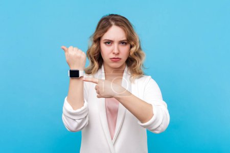 Photo for Portrait of strict bossy blond woman with wavy hair looking at camera with serious look, pointing at wristwatch, deadline, wearing white shirt. Indoor studio shot isolated on blue background. - Royalty Free Image