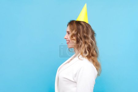 Photo for Side view portrait of joyful blond woman with wavy hair wearing white shirt and yellow party cone looking ahead with happy expression. Indoor studio shot isolated on blue background. - Royalty Free Image