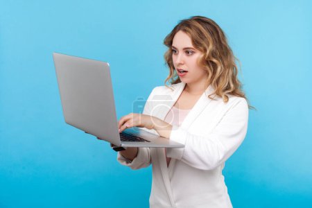 Photo for Portrait of amazed surprised blond woman freelancer with wavy hair working online on laptop looking at screen of computer, wearing white shirt. Indoor studio shot isolated on blue background. - Royalty Free Image