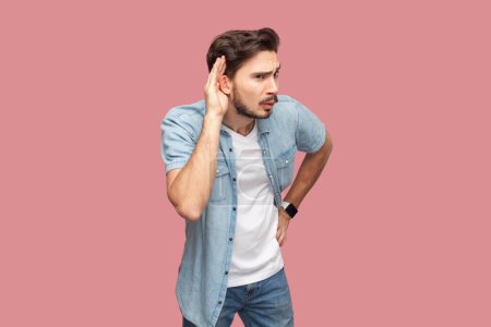 Photo for Curious bearded man standing keeps hand near ear and listens private information, tries to overhear gossiping, has intrigued expression. Indoor studio shot isolated on pink background. - Royalty Free Image
