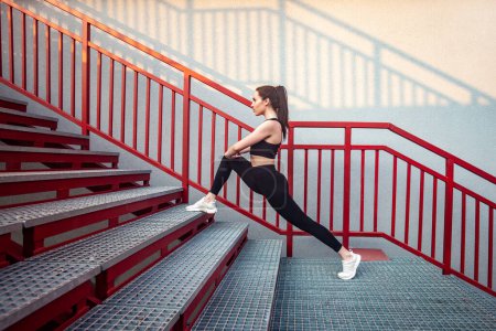 Photo for Full length portrait of slim athletic flexible woman wearing black sportswear doing physical exercising stretching legs before workout on stairs training her body outdoor. - Royalty Free Image