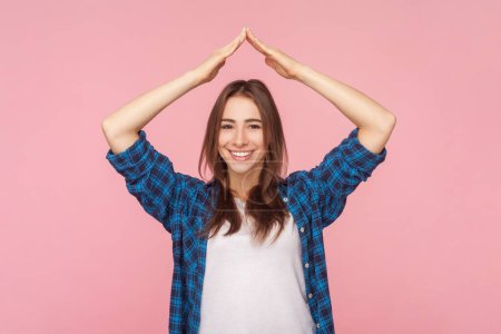 Photo for Portrait of smiling beautiful brown haired woman standing with raised arms making roof gesture feeling protection and safe, wearing checkered shirt. Indoor studio shot isolated on pink background. - Royalty Free Image