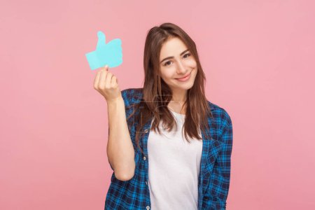 Photo for Portrait of joyful brown haired woman showing paper thumb up asking to appreciate her blog content, wearing checkered shirt. Indoor studio shot isolated on pink background. - Royalty Free Image