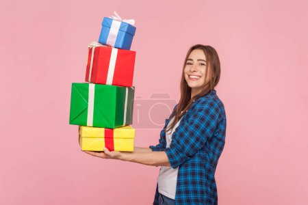 Photo for Side view portrait of satisfied brown haired woman holding lots of present boxes looking at camera enjoying birthday celebration wearing checkered shirt. Indoor studio shot isolated on pink background - Royalty Free Image