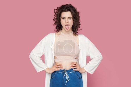 Photo for Portrait of funny childish woman with curly hairstyle wearing casual style outfit holding hands on hips, winking and sticking tongue. Indoor studio shot isolated on pink background. - Royalty Free Image