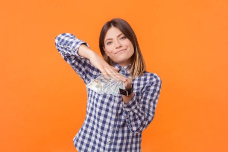 Photo for Portrait of rich confident woman with brown hair throwing dollar banknotes looking at camera winning jackpot, wearing checkered shirt. Indoor studio shot isolated on orange background - Royalty Free Image