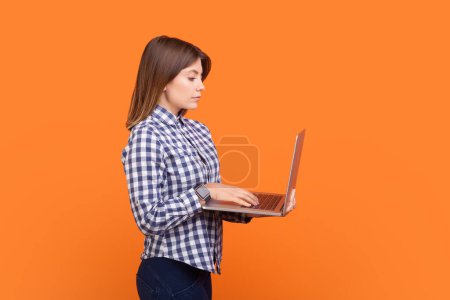 Téléchargez les photos : Portrait latéral d'une femme sérieuse confiante aux cheveux bruns travaillant sur un ordinateur portable, un projet en ligne, travaillant à la maison, portant une chemise à carreaux. Studio intérieur tourné isolé sur fond orange - en image libre de droit