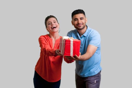 Photo for Portrait of excited optimistic couple man and woman standing together giving present box congratulating friend with holiday. Indoor studio shot isolated on gray background. - Royalty Free Image