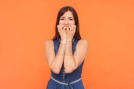 Portrait of nervous sad unhappy brunette woman wearing denim dress biting her fingernail having problems looking at camera with scared face. Indoor studio shot isolated on orange background.