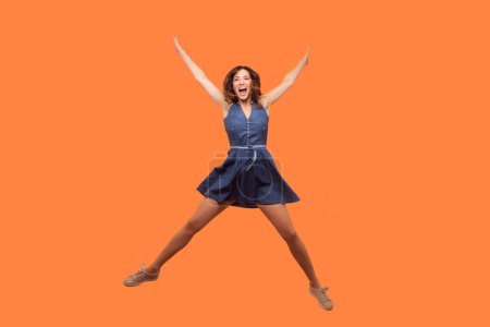 Photo for Full length portrait of funny positive happy brunette woman wearing denim dress jumping high spread hands having fun celebrating. Indoor studio shot isolated on orange background. - Royalty Free Image