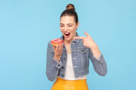 Photo for Portrait of amazed excited woman with bun hairstyle wearing denim jacket breaking diet pointing at delicious tasty donut. Indoor studio shot isolated on light blue background. - Royalty Free Image