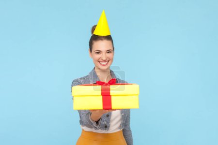 Photo for Portrait of charming beautiful friendly woman with bun hairstyle wearing denim jacket and yellow cone giving present on birthday party. Indoor studio shot isolated on light blue background. - Royalty Free Image
