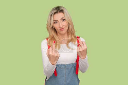 Portrait of rude impolite adult blond woman arguing with somebody, showing middle fingers, fuck gesture, wearing denim overalls. Indoor studio shot isolated on light green background