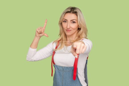 Photo for You are looser. Portrait of confident adult blond woman standing pointing to you, showing looser gesture, wearing denim overalls. Indoor studio shot isolated on light green background - Royalty Free Image