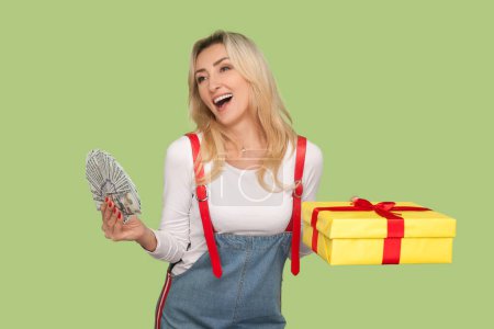 Téléchargez les photos : Portrait d'une femme blonde adulte extrêmement heureuse tenant de l'argent et une boîte cadeau, gagnant à la loterie, portant des salopettes en denim. Studio intérieur tourné isolé sur fond vert clair - en image libre de droit