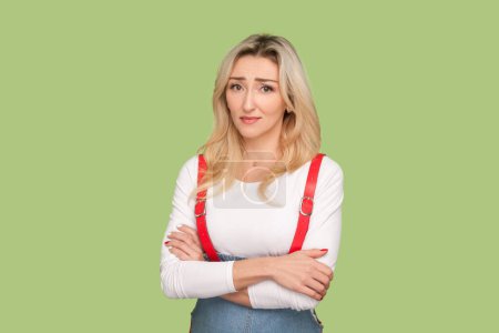 Photo for Portrait of sad depressed adult blond woman standing with folded hands, looking at camera, being offended, wearing denim overalls. Indoor studio shot isolated on light green background - Royalty Free Image