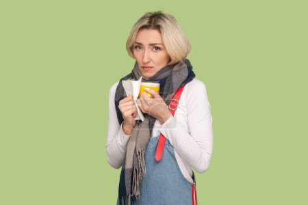 Photo for Portrait of unhealthy sick sad upset adult blond woman holding tissue and cup of tea in hands, having flu, catching cold, wearing denim overalls. Indoor studio shot isolated on light green background - Royalty Free Image