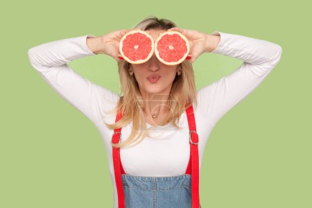 Photo for Portrait of playful funny adult blond woman standing covering her eyes with grapefruit, sending air kissing, wearing denim overalls. Indoor studio shot isolated on light green background - Royalty Free Image