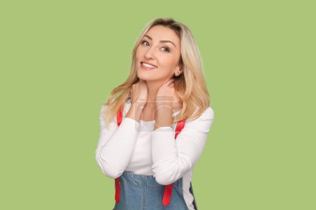 Photo for Portrait of dreaming adult blond woman looking at camera with charming smile, dreams about future vacation, wearing denim overalls. Indoor studio shot isolated on light green background - Royalty Free Image