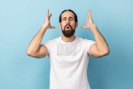 Portrait of man with beard wearing white T-shirt touching his head and showing explosion, looking worried and shocked, deadline, professional burnout. Indoor studio shot isolated on blue background.