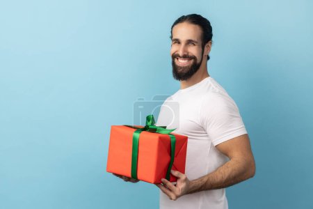 Photo for Portrait of man with beard wearing white T-shirt holding out red wrapped gift box with green ribbon, giving present, looking smiling at camera. Indoor studio shot isolated on blue background. - Royalty Free Image