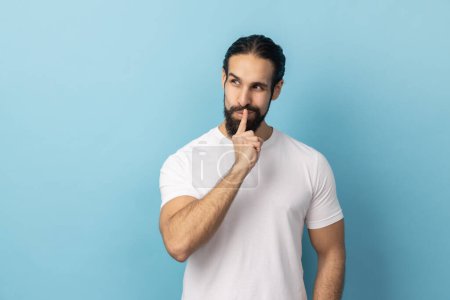 Shh, it's big secret. Portrait of man with beard wearing white T-shirt smiling, showing gesture secret sign with finger near his lips. Indoor studio shot isolated on blue background.