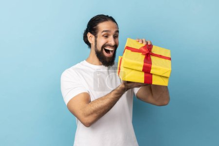 Photo for Portrait of extremely happy man with beard wearing white T-shirt looking into gift box, opening present and peeking inside with happiness. Indoor studio shot isolated on blue background. - Royalty Free Image