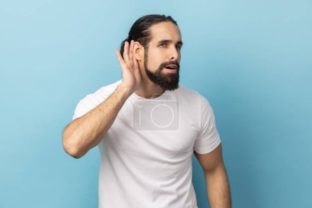 Photo for I can't hear you. Portrait of man with beard wearing white T-shirt trying to hear gossip, holding hand near ear and listening attentively carefully. Indoor studio shot isolated on blue background. - Royalty Free Image