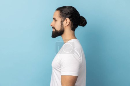Photo for Side view portrait of bearded handsome man wearing white T-shirt standing looking ahead with serious face, being strict and bossy. Indoor studio shot isolated on blue background. - Royalty Free Image