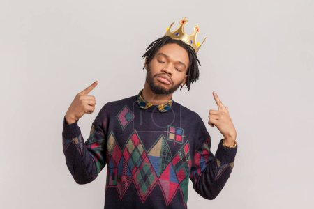 Portrait of proud self-confident handsome african-american man with dreadlocks and beard pointing at golden crown on his head. Indoor studio shot isolated on gray background.