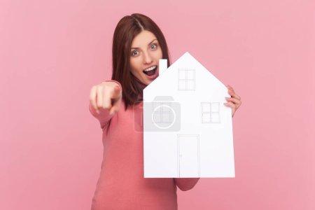 Photo for Charming woman pointing to camera and holding paper house and smiling friendly, advertising realtor services, house purchase, wearing rose turtleneck. Indoor studio shot isolated on pink background - Royalty Free Image