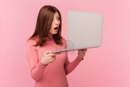 Photo for Amazed shocked woman with brown hair holding her laptop, looking at screen with big eyes and open mouth, shock content, wearing rose turtleneck. Indoor studio shot isolated on pink background - Royalty Free Image