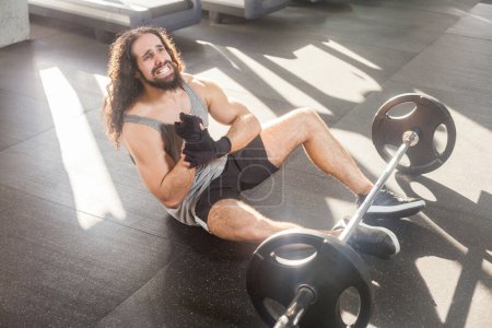 Photo for Portrait of unhealthyman with long curly hair training in gym, sitting on floor feeling strong pain in wrist, sprain in arm, frowning face and screaming, injured hand during work out. Indoor shot. - Royalty Free Image