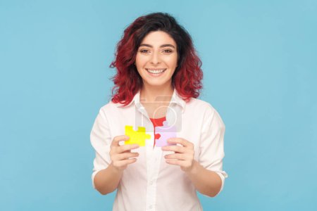 Photo for Portrait of smiling beautiful friendly woman with fancy red hair holding piece of puzzles, solving tasks, answering questions, wearing white shirt. Indoor studio shot isolated on blue background. - Royalty Free Image