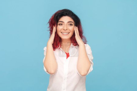 Photo for Portrait of smiling positive optimistic woman with fancy red hair dreaming about pleasant future, enjoying moment, wearing white shirt. Indoor studio shot isolated on blue background. - Royalty Free Image