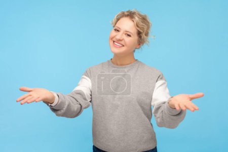 Photo for Portrait of pleased friendly smiling blonde woman saying welcome raised her arms looking at camera, wearing gray sweatshirt. Indoor studio shot isolated on blue background. - Royalty Free Image