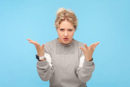 Portrait of irritated frustrated blonde woman raised her arms arguing with somebody difficult relationship, wearing gray sweatshirt. Indoor studio shot isolated on blue background.