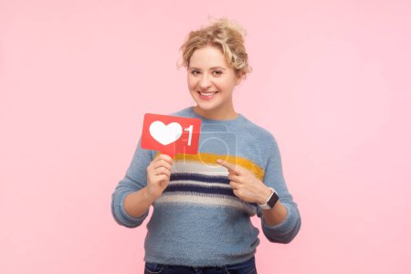 Téléchargez les photos : Portrait de sourire heureuse femme blonde positive portant sweat-shirt debout pointant vers blogueur comme icône demandant à appuyer sur le bouton. Studio intérieur isolé sur fond rose clair. - en image libre de droit