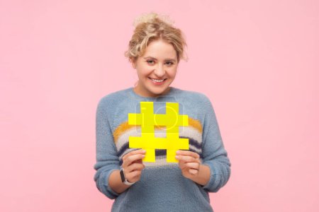 Téléchargez les photos : Portrait d'une femme blonde souriante et souriante portant un sweat-shirt portant un grand hashtag jaune regardant les tendances internet de la caméra. Studio intérieur isolé sur fond rose clair. - en image libre de droit