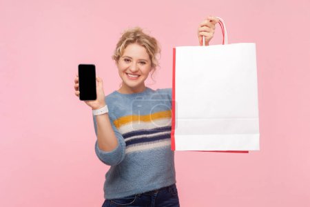 Portrait of joyful attractive blonde woman wearing sweatshirt holding shopping bags and and mobile phone with advertisement area. Indoor studio shot isolated on light pink background.